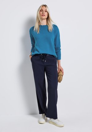 Femme blonde portant un pull bleu, un pantalon bleu marine à cordon et des baskets blanches, tenant une pochette marron, debout contre un mur blanc.