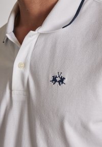 Polo bianca con accenti blu navy, caratterizzata da un colletto a due bottoni e un logo ricamato di due cavaliere. Tessuto in cotone con una trama testurizzata.