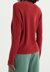 Punainen neulepaita, jonka pinnassa on tekstuuri ja ribbikuvio; siinä on rento istuvuus ja pudotetut hartiat, yhdistettynä vaaleanvihreisiin pehmeästä kankaasta valmistettuihin housuihin.