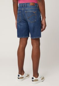 Denim shorts, donkerblauw, met gestikte accenten en een leren achterpatch. Gewerd met witte en beige sneakers.