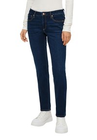 Jeans slim-fit bleu marine en denim, dotés de cinq poches, d'une fermeture à bouton et de coutures contrastées. Portés avec des sneakers blanches à semelles épaisses.