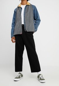 Personne portant une veste en jean avec un col beige, une chemise blanche, une écharpe grise, un pantalon noir, des chaussettes blanches et des baskets vertes, debout devant un fond uni.