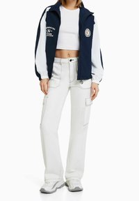 Coupe-vent bleu marine et blanc avec fermeture éclair à l'avant, poignets élastiques et accents graphiques, associé à un pantalon cargo blanc taille haute avec poches.