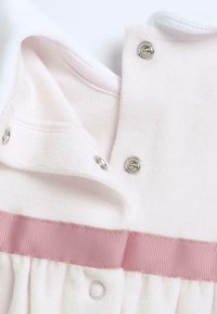 Svetle růžový oděv s jemnou texturou, zdobený saténovou stuhou a patentkami u výstřihu. Obsahuje detail zaobleného límce.