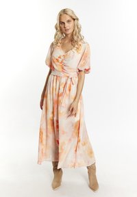 Abito maxi floreale in un delicato color pesca, con scollatura a V, maniche corte a palloncino, dettagli con bottoni e vita con fiocco. Indossato con stivaletti beige alla caviglia.