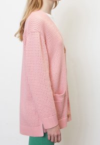 Růžový pletený kardigan s volným střihem, širokými rukávy a texturou ve tvaru včelí plástve; obsahuje dvě kapsy a zapínání na knoflík.