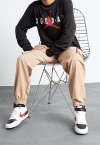 Svart långärmad tröja med röd grafik, ljusbruna byxor med elastiska muddar och vita/röda Nike högklackade sneakers. Metallstol i bakgrunden.