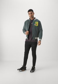 Giacca bomber in corduroy verde con un patch a forma di lettera "B" giallo e rosso, abbinata a pantaloni neri e sneakers nero e oro.
