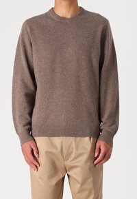 Brązowy sweter z okrągłym dekoltem wykonany z miękkiego materiału, z prążkowanymi mankietami i dołem, noszony na jasnobeżowych spodniach.