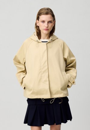 Chaqueta con capucha beige de corte relajado, con mangas raglán, dos bolsillos y detalles de cordón. Llevada sobre una falda de tela con pliegues en azul marino.