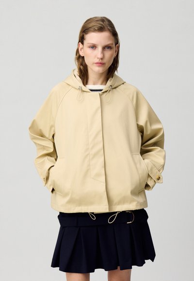 Veste à capuche beige avec une coupe décontractée, présentant des manches raglan, deux poches et des détails à cordon. Portée par-dessus une jupe plissée bleu marine.