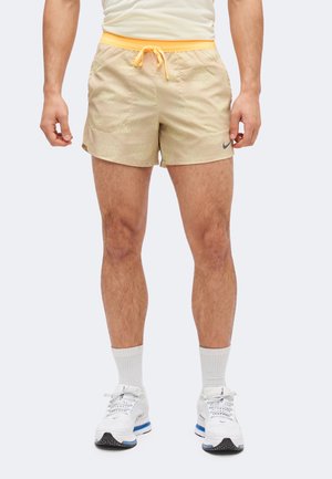 Άνδρας που φοράει μπεζ σορτς με κίτρινη μέση και κορδόνι, λευκές κάλτσες και λευκά αθλητικά παπούτσια, στέκεται μπροστά σε λευκό φόντο.