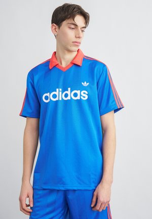 Νεαρός άνδρας που φοράει μπλε αθλητική φανέλα Adidas με κόκκινο γιακά και ρίγες, κοιτάζοντας ελαφρώς προς τα κάτω και πλάγια.