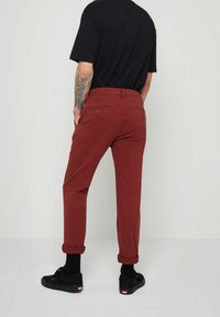 Homme portant un pantalon roux retroussé, un t-shirt noir, des chaussettes noires et des baskets noires, debout avec les mains dans les poches arrière.