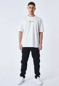 Camiseta blanca de gran tamaño con un diseño gráfico en el pecho, combinada con pantalones cargo negros que cuentan con múltiples bolsillos y zapatillas.
