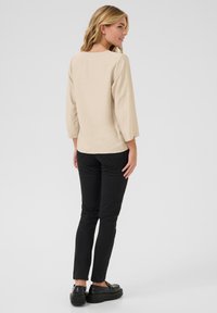 Haut beige à manches longues avec un col rond, associé à un pantalon noir slim-fit et des chaussures noires. Le haut présente un tissu texturé et des manches trois-quarts.