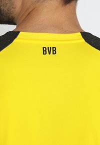 Gul tröja med svarta accenter och korta ärmar. Har ett texturerat tyg och "BVB"-logotypen tryckt i svart på ryggen.