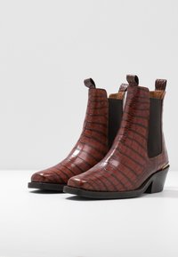 Des bottes de cheville en cuir brun embossées crocodile avec des panneaux latéraux élastiques et un petit talon, mises en valeur sur un fond blanc uni.