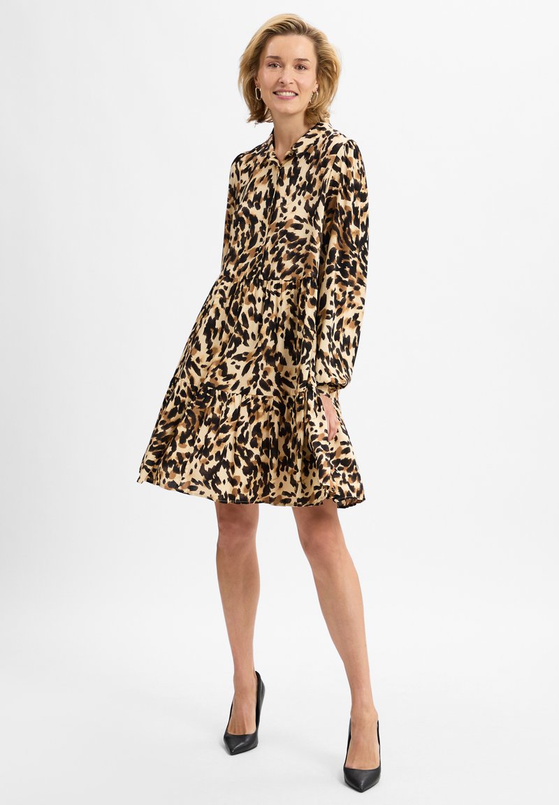 Tiered Kleid mit Tierdruck in Beige und Schwarz, ausgestattet mit einer Knopfleiste, Kragen, langen Ärmeln und einem schwingenden, gestuften Saum. Kombiniert mit schwarzen Absätzen.