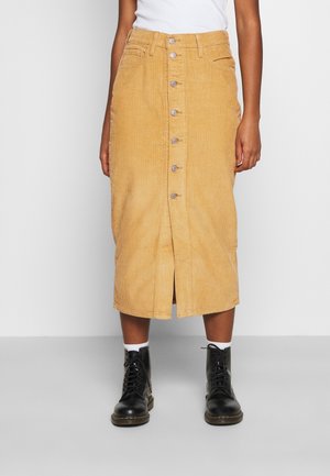 Femme portant une jupe midi en velours côtelé couleur camel avec des boutons sur le devant, des bottes noires à lacets, des chaussettes blanches et un haut blanc, debout sur un sol gris.