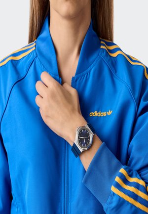 Persona, kas valkā zilu Adidas jaku ar dzeltenām svītrām, rāda sudraba pulksteni ar zilu siksniņu uz kreisā plaukstas.