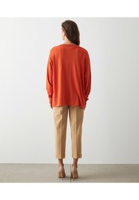 Przewymiarowany pomarańczowy sweter z dzianiny z długimi rękawami, zestawiony z beżowymi spodenkami o długości do kostek i cielistymi szpilkami. Minimalistyczne tło.