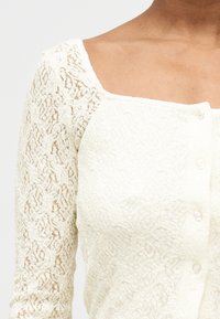 Κοντινό πλάνο ενός ατόμου που φοράει εκρού δαντελένιο ζακέτα με λουλ ουδάτα σχέδια και μικρά λευκά κουμπιά στο μπροστινό μέρος.