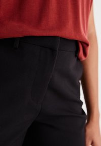 Zwarte op maat gemaakte broek met een gladde textuur en subtiele zijzak, gecombineerd met een gedrapeerde rode top die de tailleband accentueert.