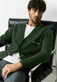 Groene gebreide cardigan met een V-hals, knoopsluiting en geribbelde manchetten. Heeft witte geborduurde tekst aan de linkerkant. Gecombineerd met een wit T-shirt.
