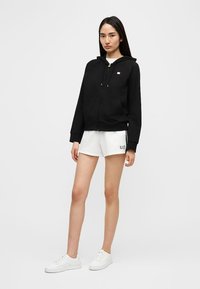 Μαύρο φούτερ με φερμουάρ και μπροστινή τσέπη, με μικρό λογότυπο. Συνδυασμένο με άσπρες σορτς και λευκά αθλητικά παπούτσια, σε ουδέτερο φόντο.