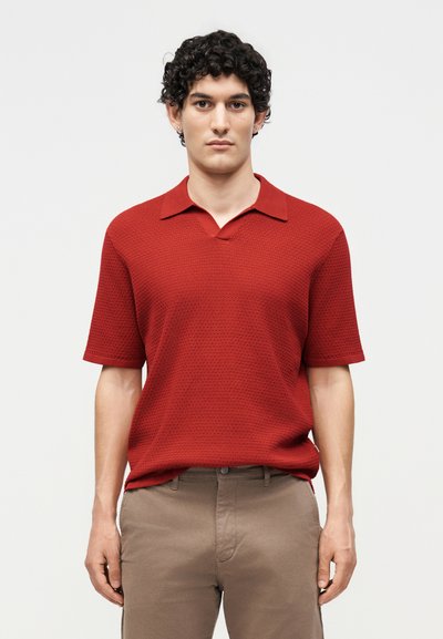 Homme aux cheveux bouclés foncés, portant un polo rouge texturé à manches courtes et un pantalon beige, debout devant un fond clair uni.