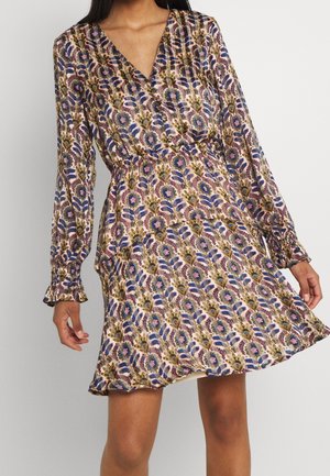 Femme portant une robe à manches longues et col en V avec une taille élastique, motif floral aux tons violets, bleus, verts et beiges, debout devant un fond uni.