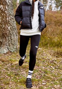 Chaqueta de abrigo negra, sudadera blanca con capucha, y leggings negros con logo. Zapatos negros con calcetines rayados blancos. Entorno al aire libre con hierba y árboles.