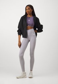 Čierna mikina na zips, fialový crop top s logom, svetlošedé legíny s vysokým pásom a biele atletické topánky. Hladká tkanina a priliehavý strih.