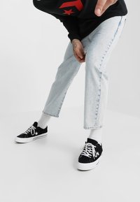 Person in hellblauen, verkürzten Jeans, weißen Socken und schwarzen Sneakers mit weißen Schnürsenkeln, die sich leicht nach vorne beugt vor einem einfarbigen Hintergrund.