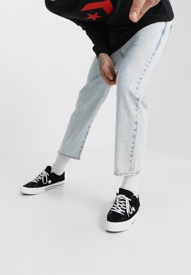 Person in hellblauen, verkürzten Jeans, weißen Socken und schwarzen Sneakers mit weißen Schnürsenkeln, die sich leicht nach vorne beugt vor einem einfarbigen Hintergrund.