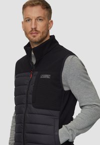 Schwarze Daunenweste mit hohem Kragen, ausgestattet mit einem Reißverschluss, einer linken Brusttasche und gestepptem Muster. Graues Langarmshirt darunter.
