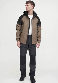 Jachetă waterproof maro și neagră, cu glugă, buzunar pe piept cu fermoar și manșete ajustabile, asociată cu pantaloni închiși la culoare cu petice la genunchi.