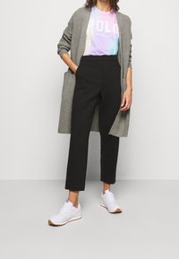 Kvinna i en rosa Polo Ralph Lauren skjorta, grå cardigan, svarta cropped byxor och vita sneakers, stående med ena handen i fickan.