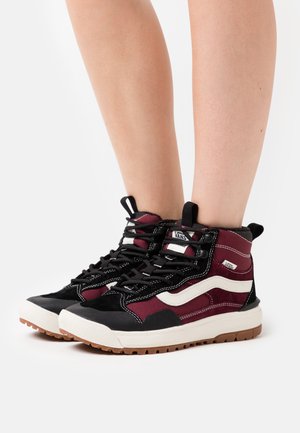 Beine, die schwarze, bordeauxrote und weiße High-Top-Vans-Sneaker mit dicken weißen Sohlen und braunem Profil tragen, stehend auf weißem Hintergrund.