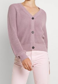 Kvinna klädd i en mauve-färgad stickad cardigan med svarta knappar och ljusrosa byxor, avskuren vid halsen och höfterna.