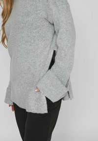 Hellgrauer Langarmpullover mit strukturiertem Strick, seitlichen Schlitzen und entspannter Passform, kombiniert mit schwarzen Hosen.