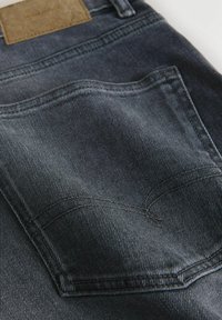 Bolsillo trasero de mezclilla gris oscuro con costura curva doble en tela de jeans y una etiqueta de cuero marrón en la cintura.