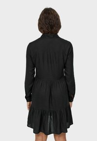 Svart långärmad klänning med figurnära överdel och volangkjol. Tillverkad av ett lätt tyg, med krage och knappade ärmslut.