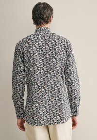 Blumiges Hemd aus dunklem Stoff mit kleinen weißen und blauen Blumen. Mit langen Ärmeln und geradem Schnitt sowie einem Rückenkraus.