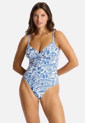 Donna che indossa un costume intero blu e bianco con fantasia e spalline sottili, in piedi con le mani dietro la schiena su uno sfondo semplice.