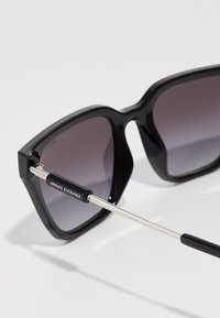 Ochelari de soare supradimensionați, de culoare neagră, cu lentile degradate, având un cadru din plastic lucios și sicră metalică argintie. Logo-ul este accentuat pe sicră.