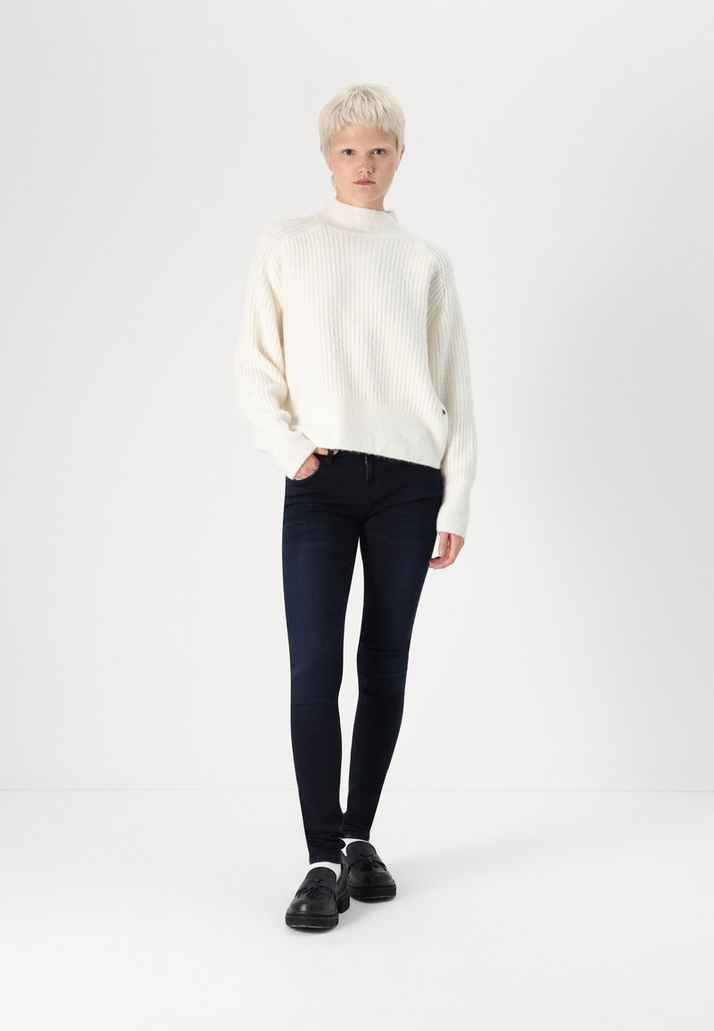 Maglione bianco a coste, jeans skinny scuri e scarpe nere. Il maglione presenta un collo alto e una vestibilità ampia, creando un aspetto accogliente.