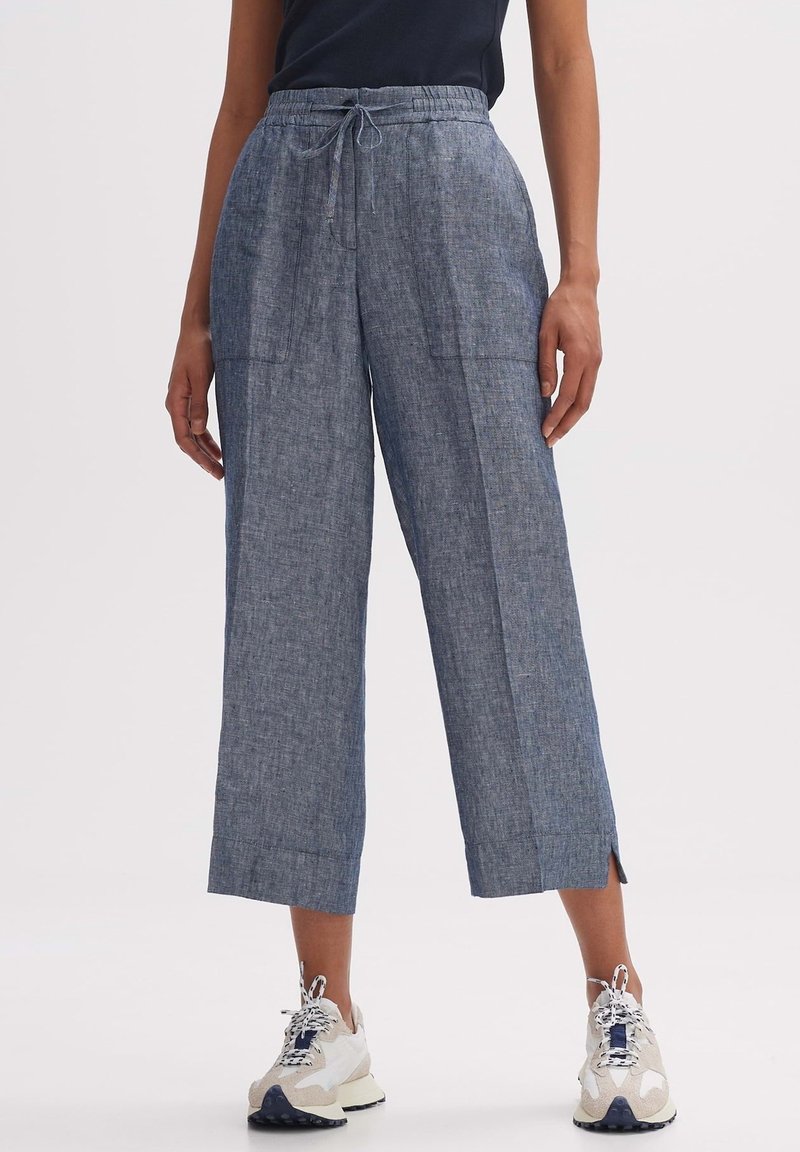 Blue linen culotte pants with a drawstring waist, side pockets, and slits near the hem. Paired with beige and white sneakers.