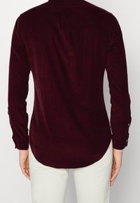 Camisa de pana burdeos de manga larga, con puños de botón y dobladillo redondeado. Textura suave y patrón de canalé sutil, vista desde la parte trasera.